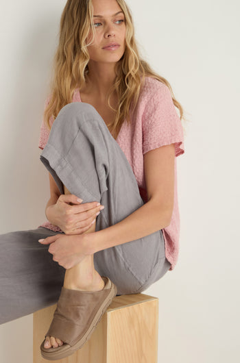 A woman with long blond hair sits on a wooden block, wearing a textured pink top, gray pants, and tan Crushed Leather Sliders, looking off to the side.