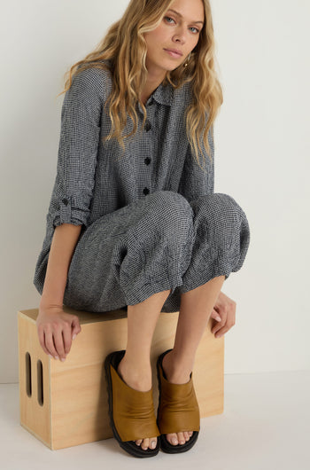 A woman in a gray checkered outfit sits on a wooden stool with one leg up, wearing stylish Crushed Leather Sliders, against a plain light background.