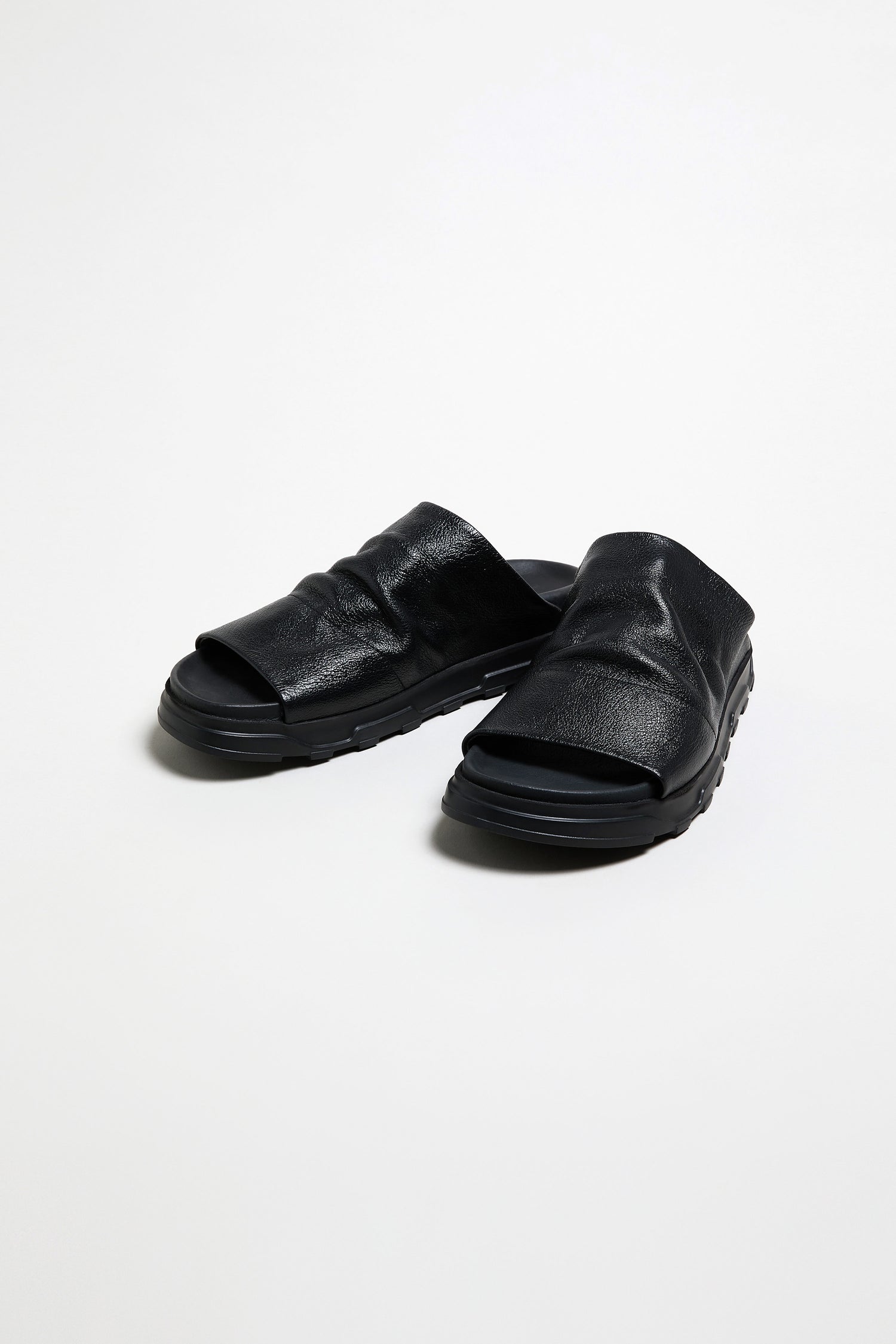 The Crushed Metallic Leather Sliders, handcrafted in Italy with thick soles, are luxury slip-on sandals in black leather, displayed on a plain white background.