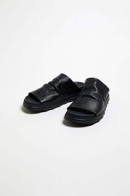The Crushed Metallic Leather Sliders, handcrafted in Italy with thick soles, are luxury slip-on sandals in black leather, displayed on a plain white background.