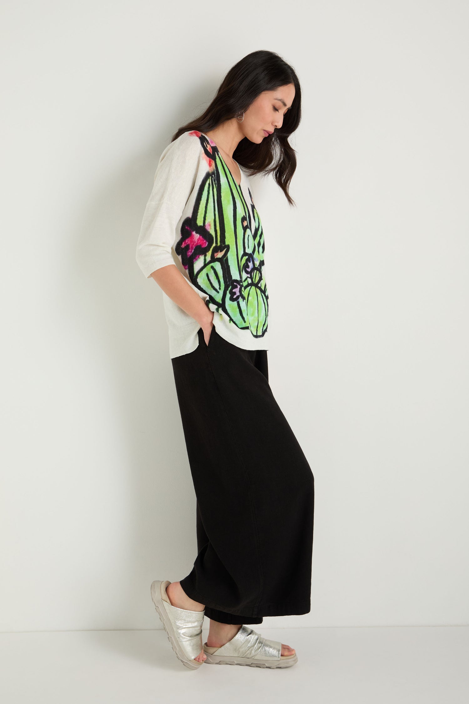 A woman stands against a white wall, wearing a white top with a colorful abstract cactus design, black wide-leg pants, and Crushed Metallic Leather Sliders handcrafted in Italy.