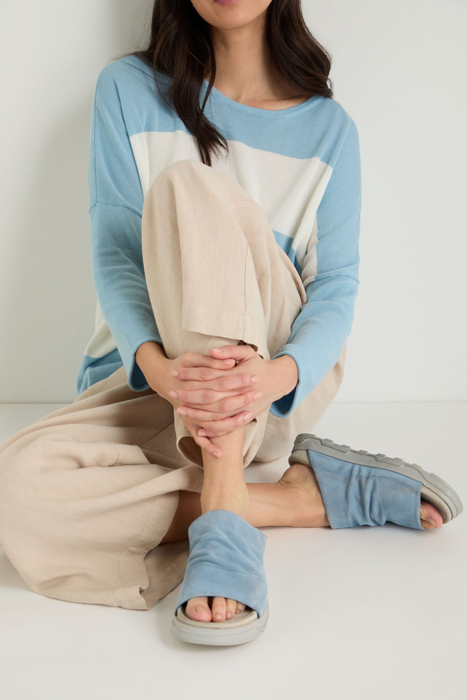 A woman sits cross-legged on the floor, wearing a blue and white top, beige wide-leg pants, and blue Crushed Suede Sliders by Lofina—crafted in Italy.