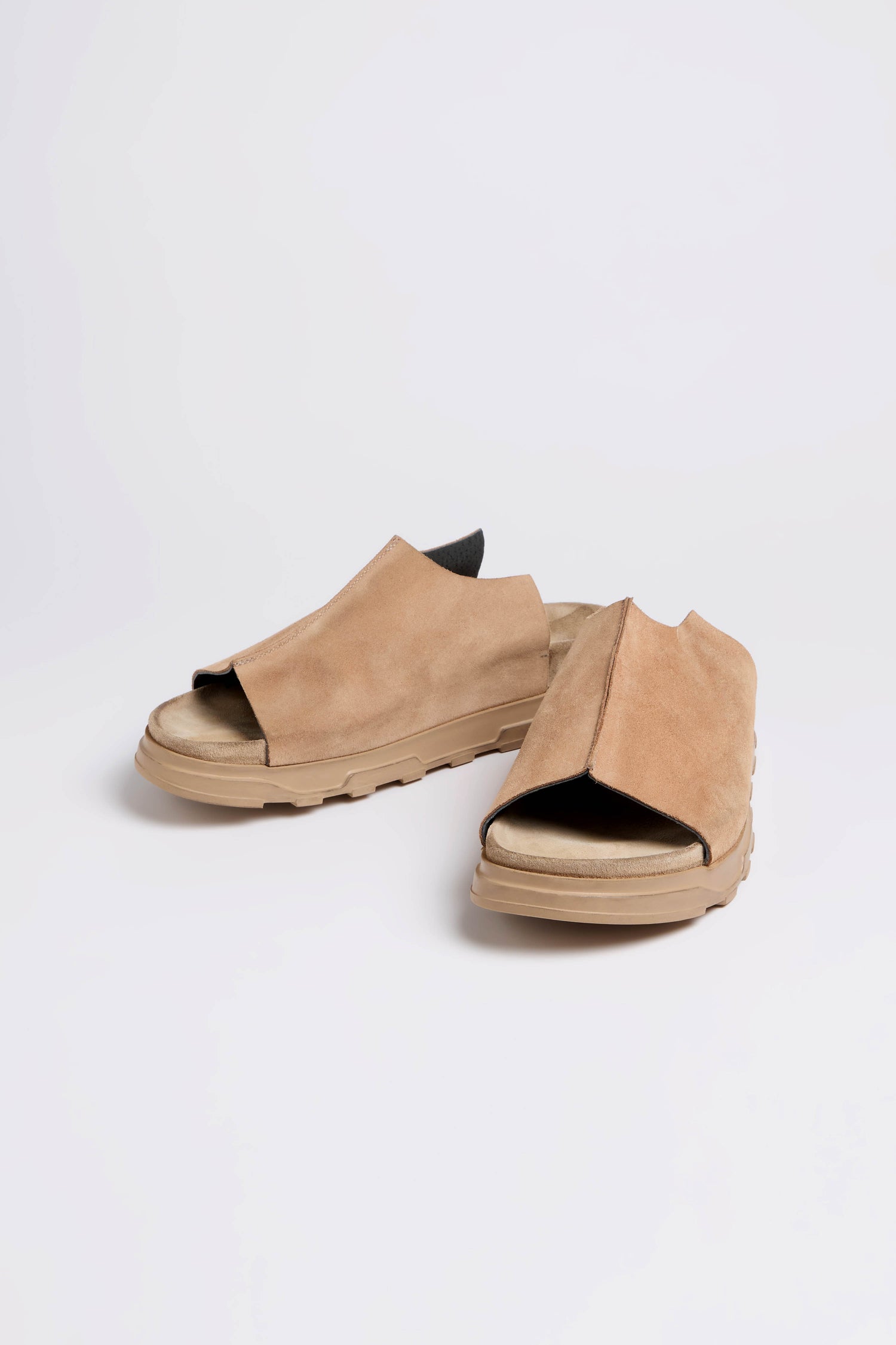 The Seam Suede Sliders, featuring brown suede with open toes and chunky tan soles, are showcased on a white background. Handcrafted in Italy, these sliders perfectly merge contemporary minimalism with style and comfort.