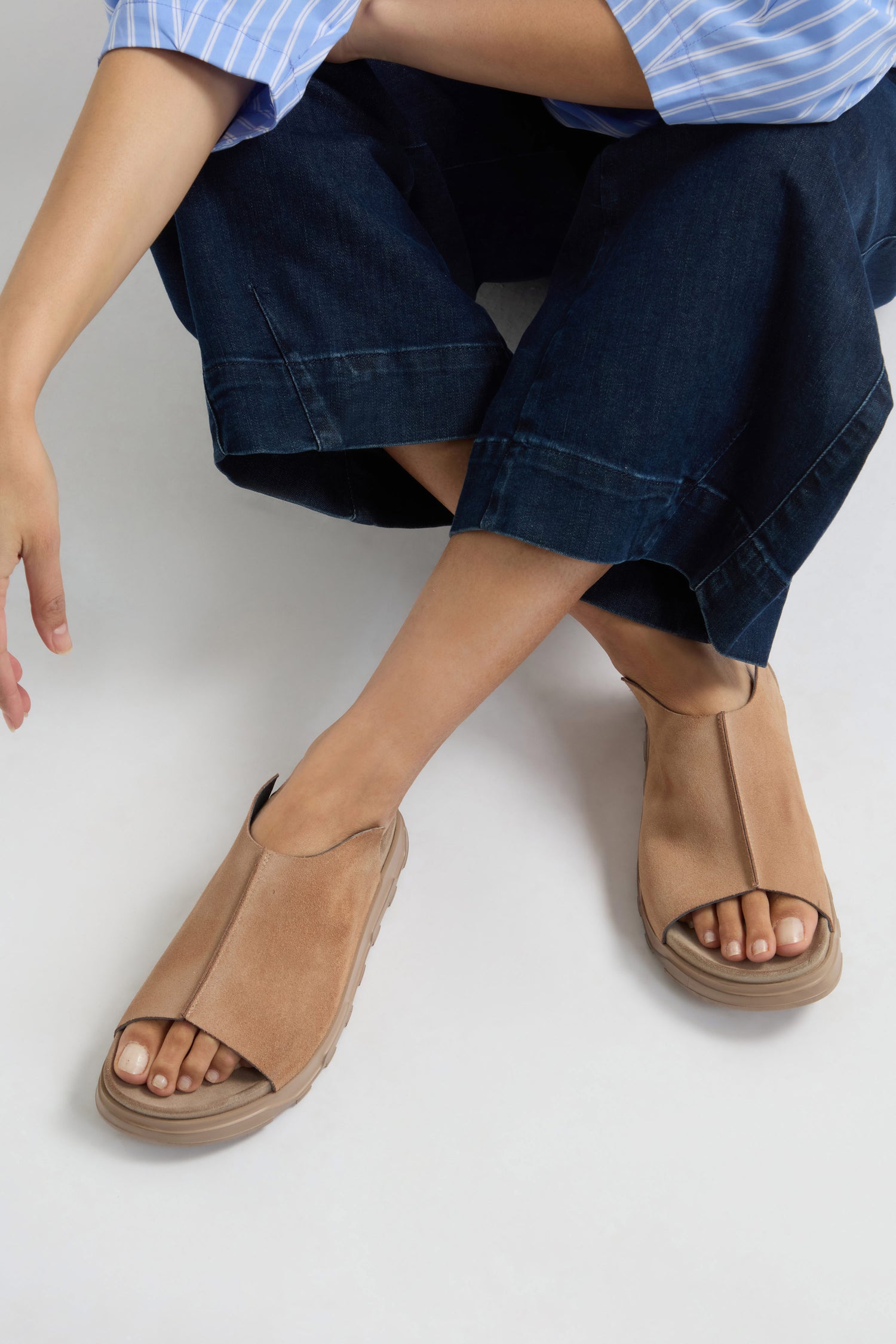 A person embodies contemporary minimalism while seated on a white surface, sporting dark wide-leg pants paired with tan Seam Suede Sliders.