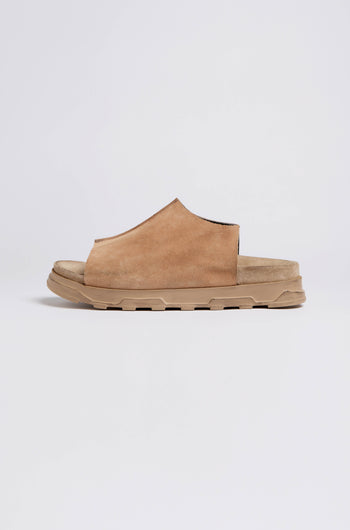 A side view of the Seam Suede Sliders, a brown suede slip-on sandal showcasing contemporary minimalism. It features a textured beige rubber sole and is handcrafted in Italy, set against a plain white background.