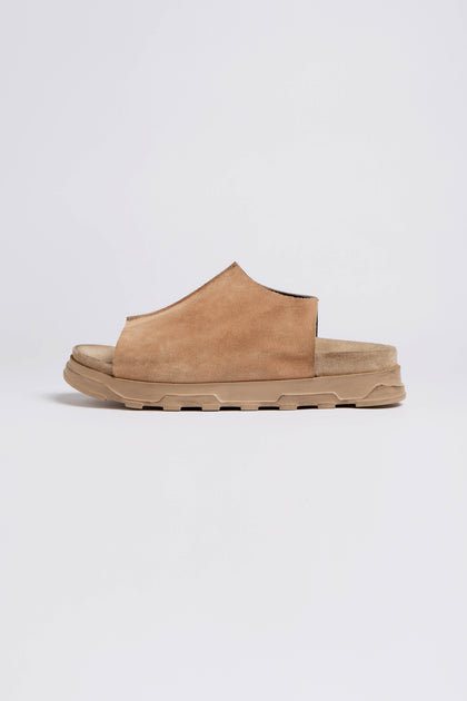 A side view of the Seam Suede Sliders, a brown suede slip-on sandal showcasing contemporary minimalism. It features a textured beige rubber sole and is handcrafted in Italy, set against a plain white background.