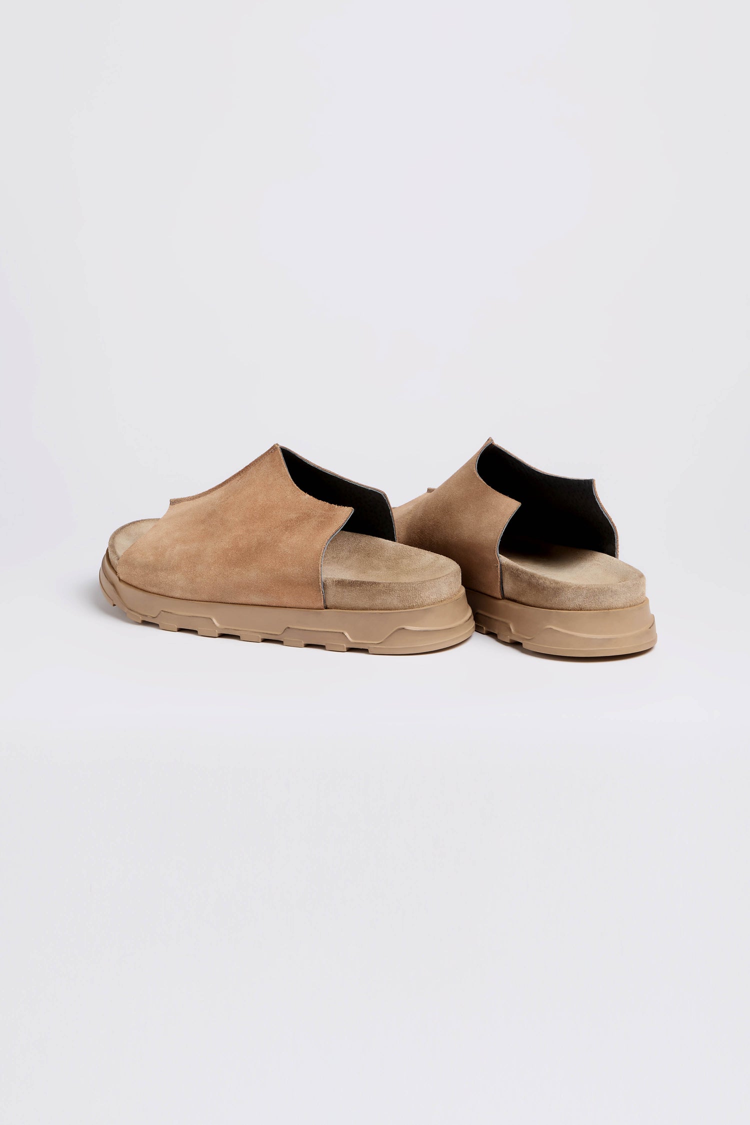 The Seam Suede Sliders, handcrafted in Italy, offer contemporary minimalism with their brown slip-on design and chunky rubber sole. Viewed from the back and side against a plain background, these shoes blend style and quality seamlessly.