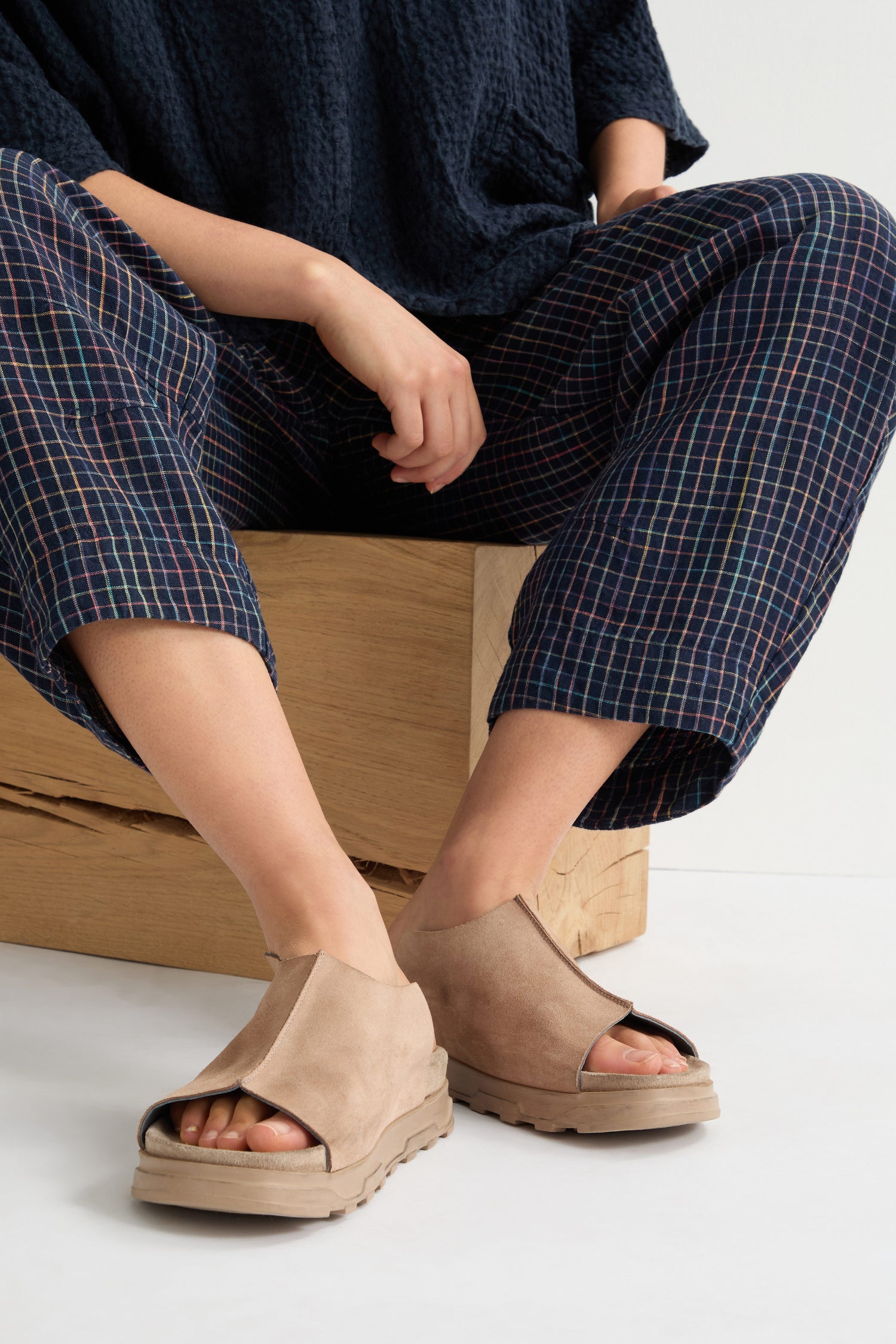 Seated with legs crossed, a person showcases contemporary minimalism in tan Seam Suede Sliders and dark blue wide-leg pants.