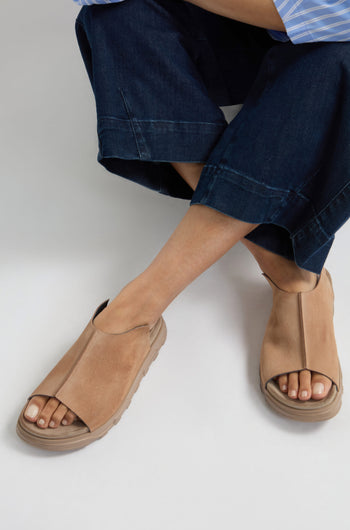 Seated with legs crossed, a person showcases contemporary minimalism in tan Seam Suede Sliders and dark blue wide-leg pants.