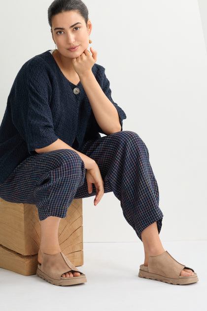 A seated person embodies contemporary minimalism wearing a dark shirt, plaid pants, and Seam Suede Sliders. They pose confidently on a wooden cube against a pristine white background, showcasing Italian craftsmanship.