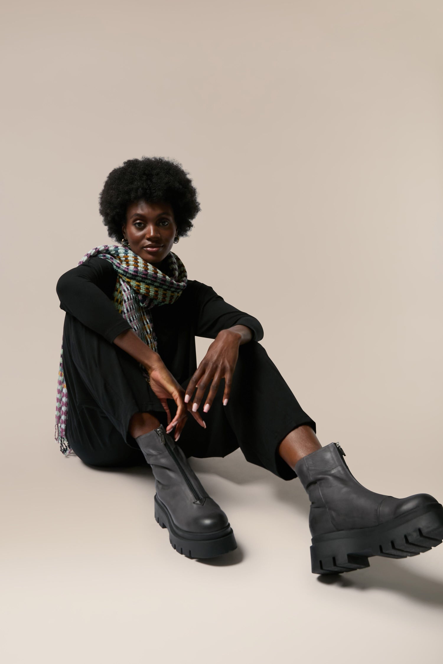 A black woman sitting on a grey background in a scarf and black Platform Zip Front Leather Boots.