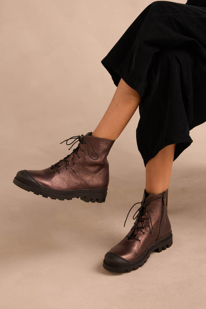 Person wearing black pants and Metallic Chocolate Lace Up Leather Boots, crafted in calfskin with thick rubber soles, seated against a neutral background.