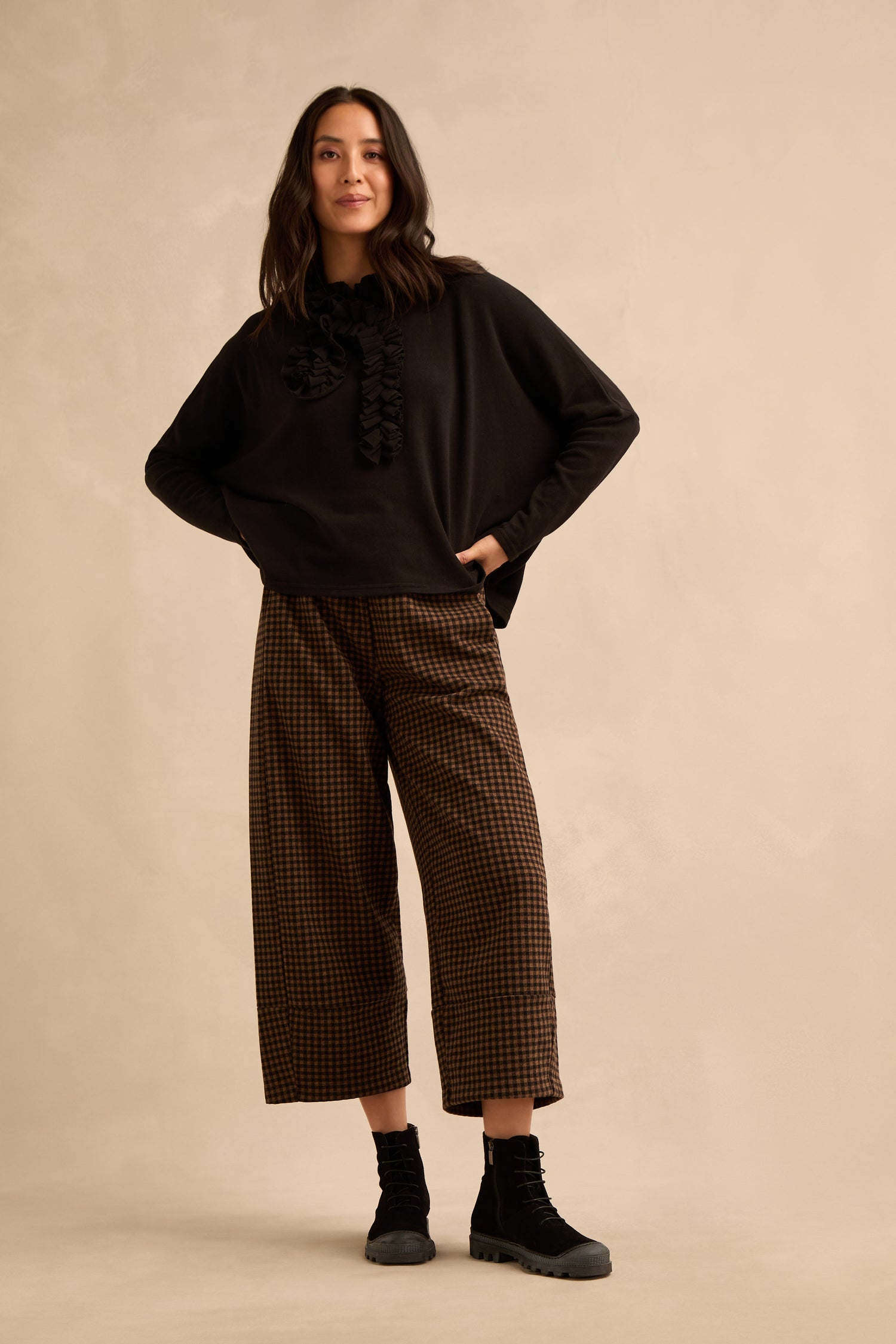 A woman stands against a beige background, wearing a black long-sleeve top with ruffle detail, brown checkered wide-leg pants, and Lace Up Suede Boots.