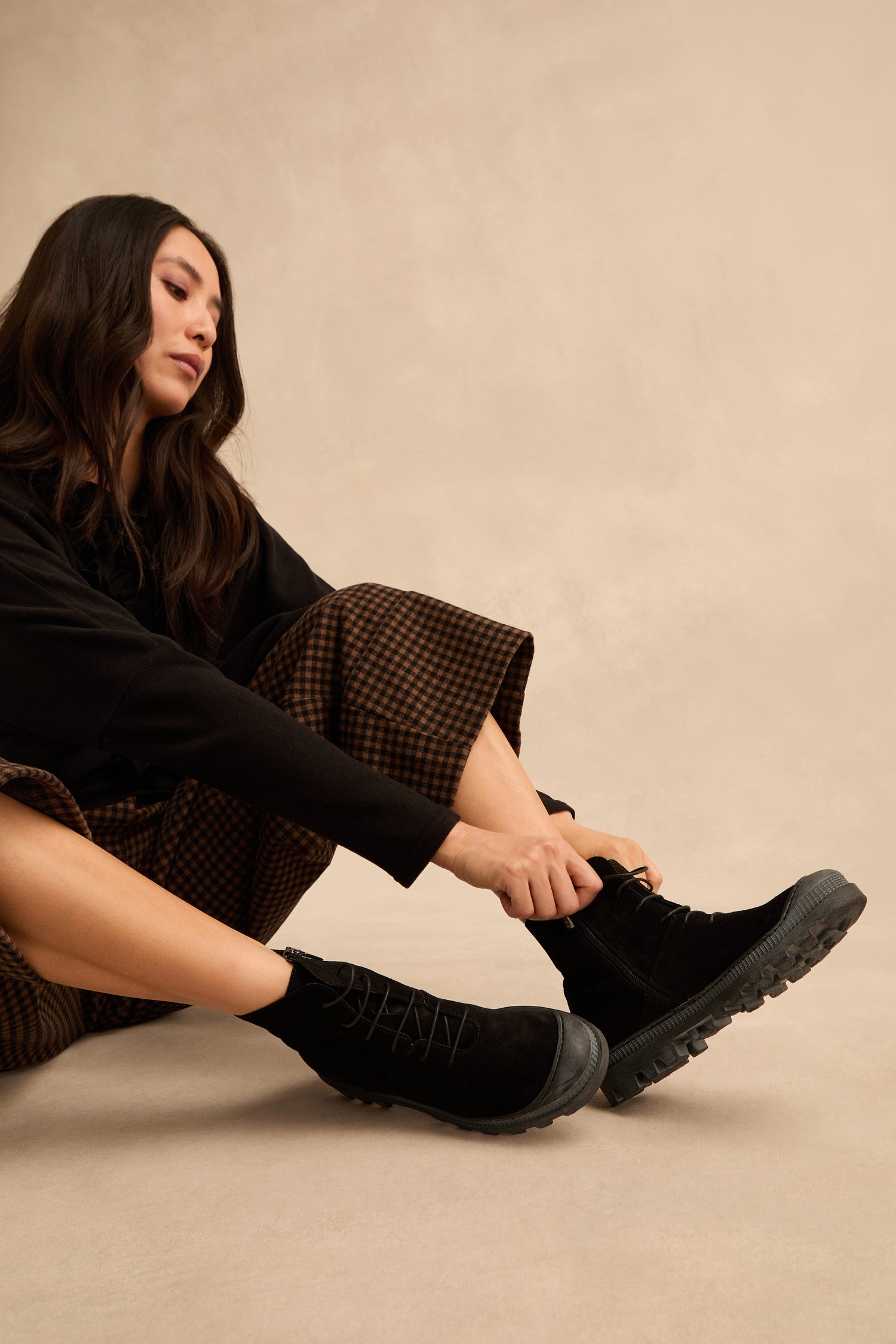 Seated on the floor against a neutral background, a person wearing a black top and brown checkered pants puts on Lace Up Suede Boots.