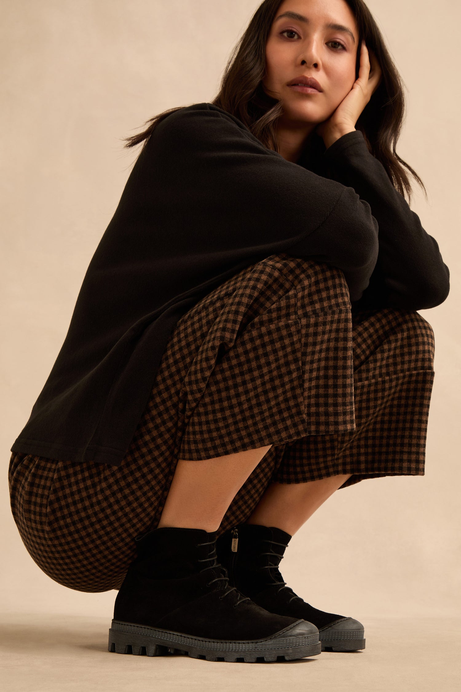 A woman squats against a plain background, wearing a black long-sleeve top, brown checkered pants, and Lace Up Suede Boots, with one hand resting on her face.