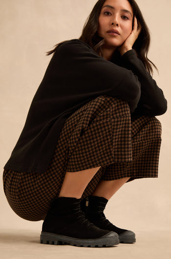 A woman squats against a plain background, wearing a black long-sleeve top, brown checkered pants, and Lace Up Suede Boots, with one hand resting on her face.