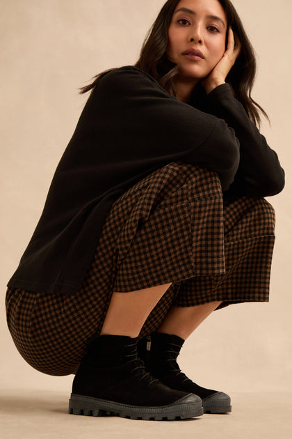 A woman squats against a plain background, wearing a black long-sleeve top, brown checkered pants, and Lace Up Suede Boots, with one hand resting on her face.