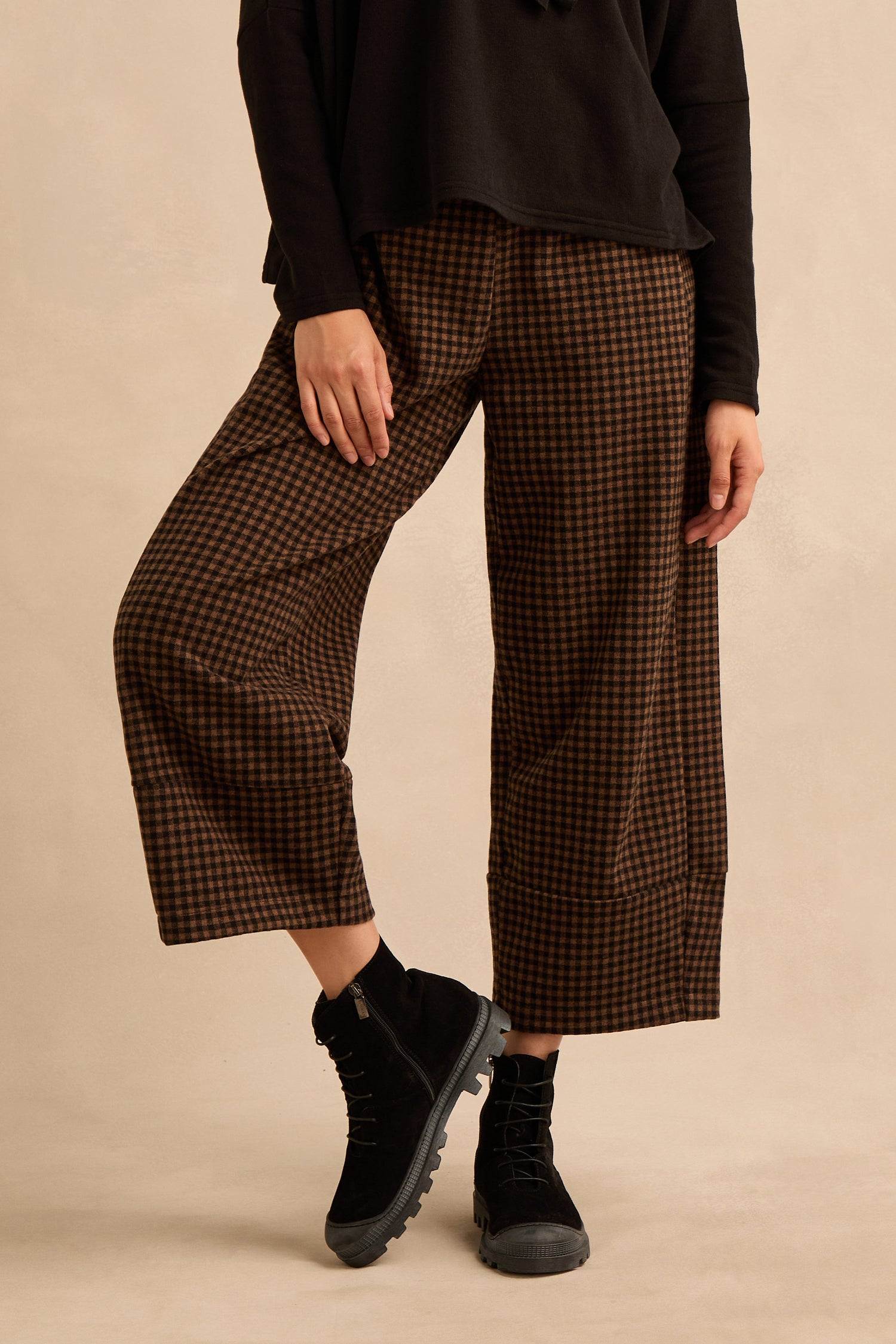 Wearing brown checkered wide-leg pants, a black long-sleeve top, and Lace Up Suede Boots, the person stands against a plain beige background.