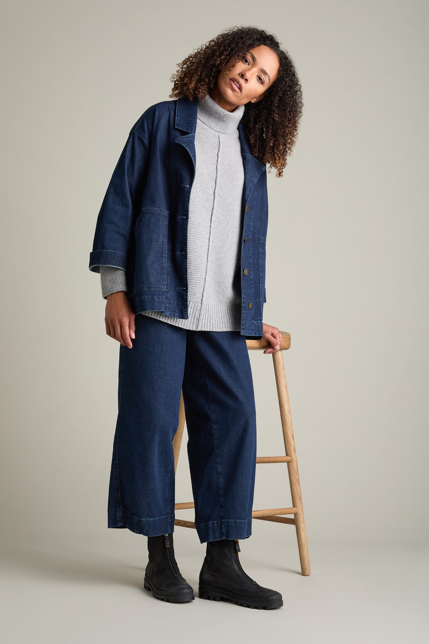 A woman with curly hair, wearing a grey turtleneck sweater, blue denim jacket, and matching wide-leg pants, leans against a wooden stool in a neutral-toned room. She completes her stylish outfit with Zip Front Leather Boots in soft calfskin.