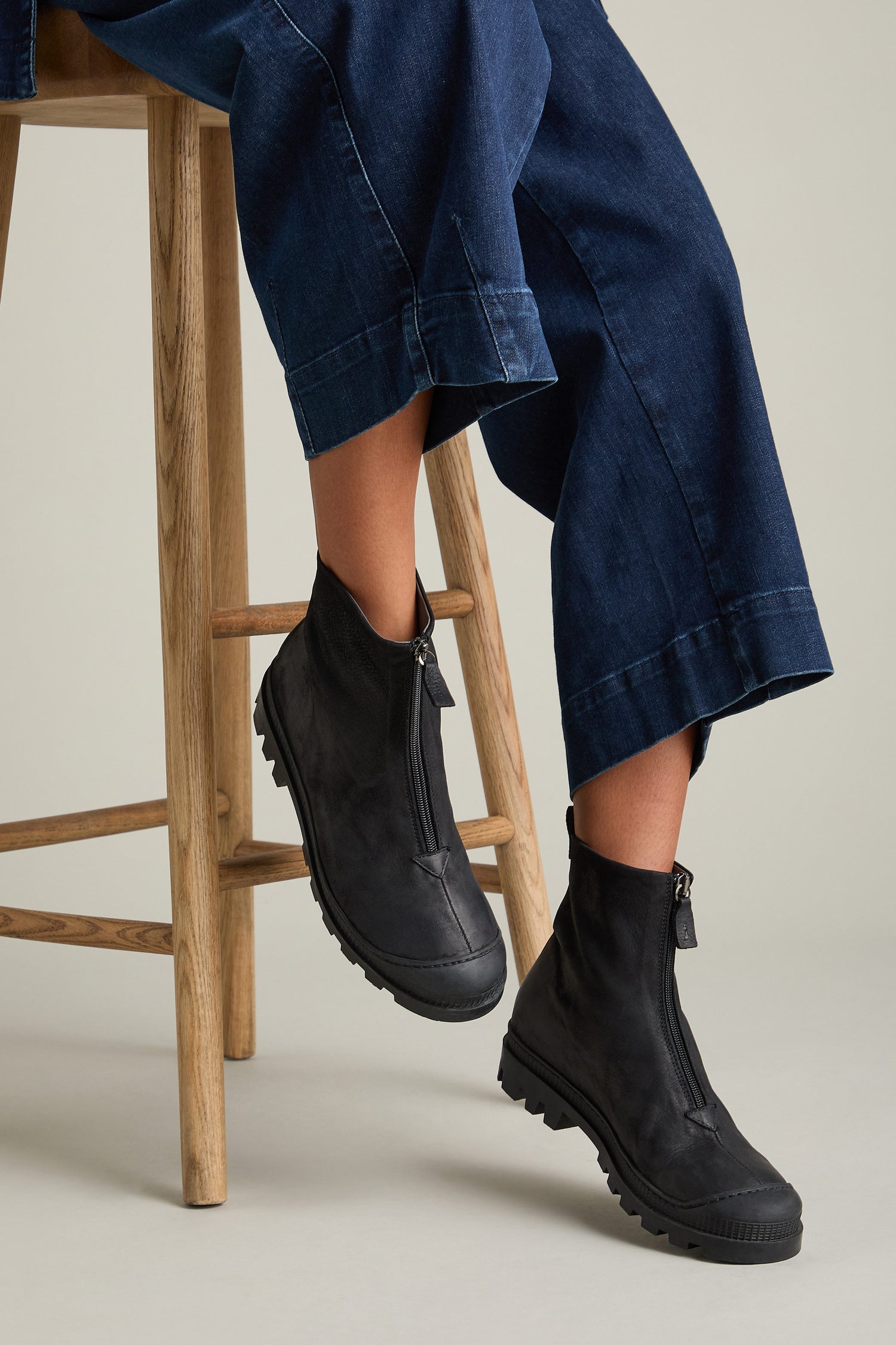 Person sitting on a wooden stool, wearing dark blue wide-leg jeans and black Zip Front Leather Boots from Lofina crafted in luxurious calfskin.