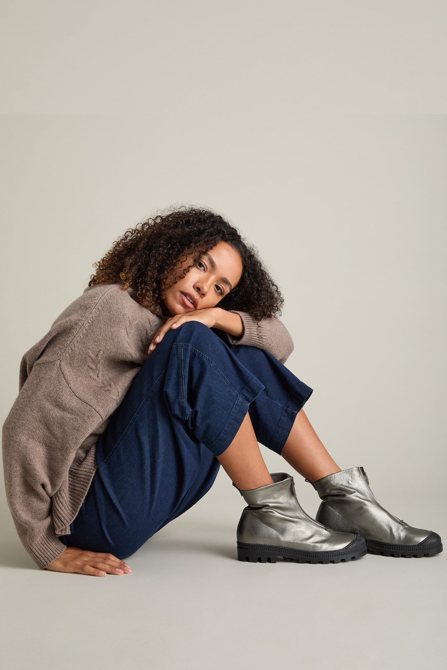 A person with curly hair sits on the floor, wearing a brown sweater, blue pants, and metallic silver Zip Front Leather Boots.