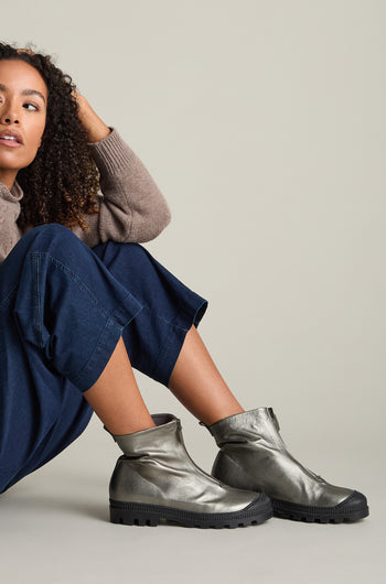 A person in a brown sweater and blue pants sits on the floor, wearing Zip Front Leather Boots in metallic gray crafted from calfskin with black soles.