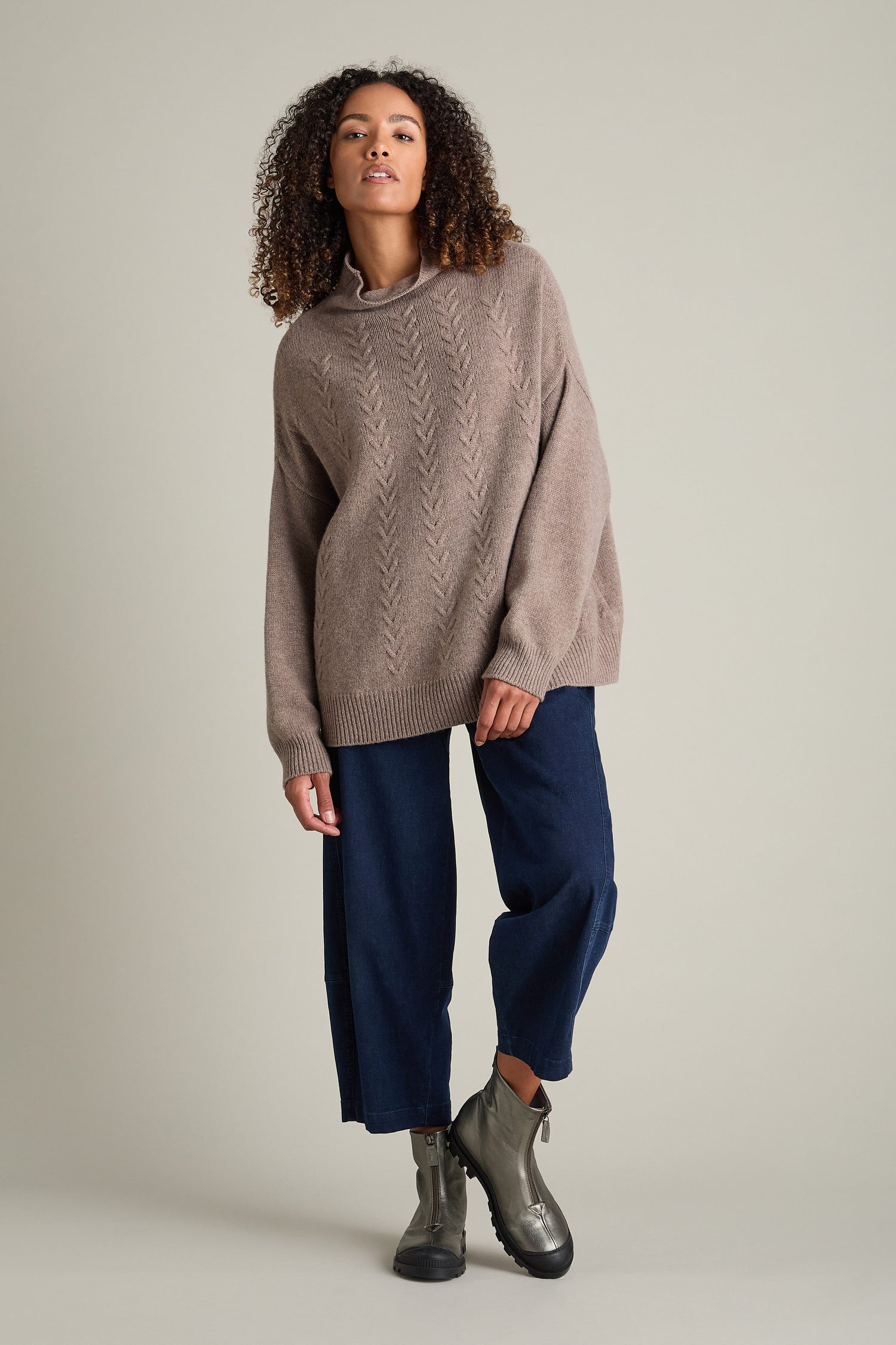 A person with curly hair is wearing a brown knit sweater, dark blue pants, and Zip Front Leather Boots from Lofina, posing against a plain grey background.