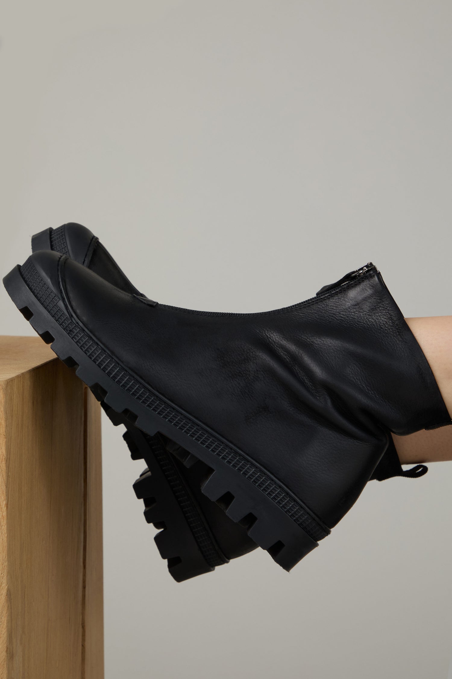 A person wearing Black Zip Front Leather Boots with thick, treaded soles rests their feet on a light wooden surface against a plain background.