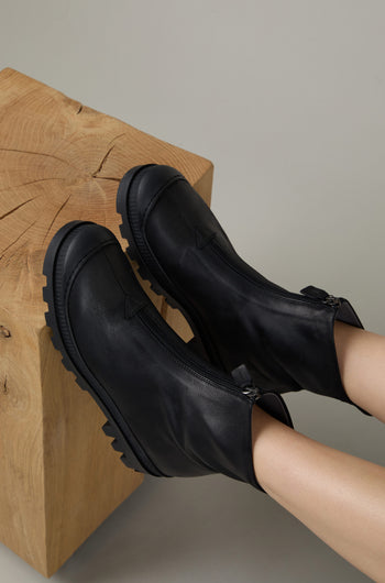Two feet in Black Zip Front Leather Boots with chunky soles rest on a light wooden cube with visible grain patterns.