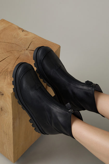Two feet in Black Zip Front Leather Boots with chunky soles rest on a light wooden cube with visible grain patterns.