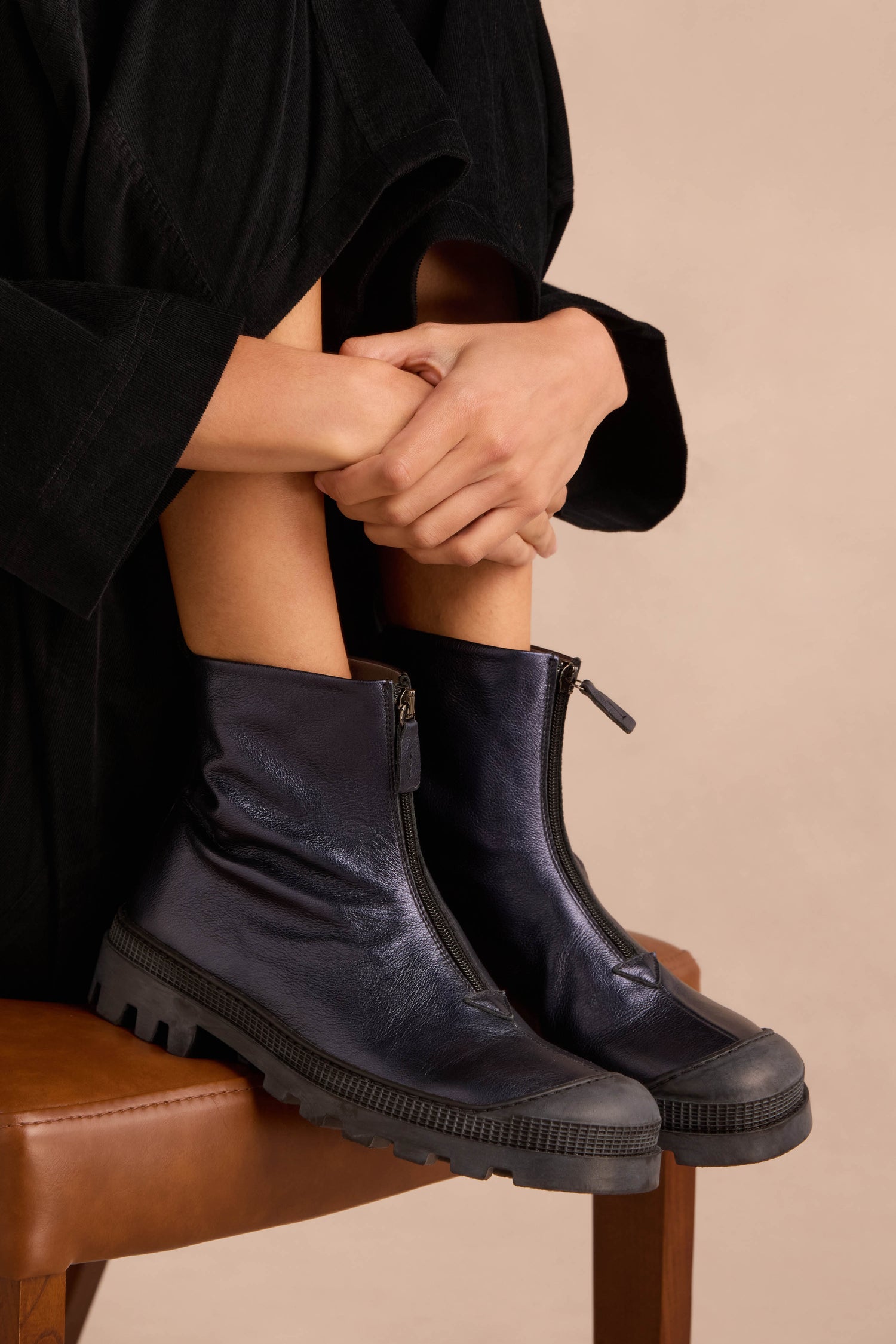 A person in a black outfit sits on a brown chair, hugging their knees and wearing Metallic Blue Violet Zip Front Leather Boots with a chunky tread sole.