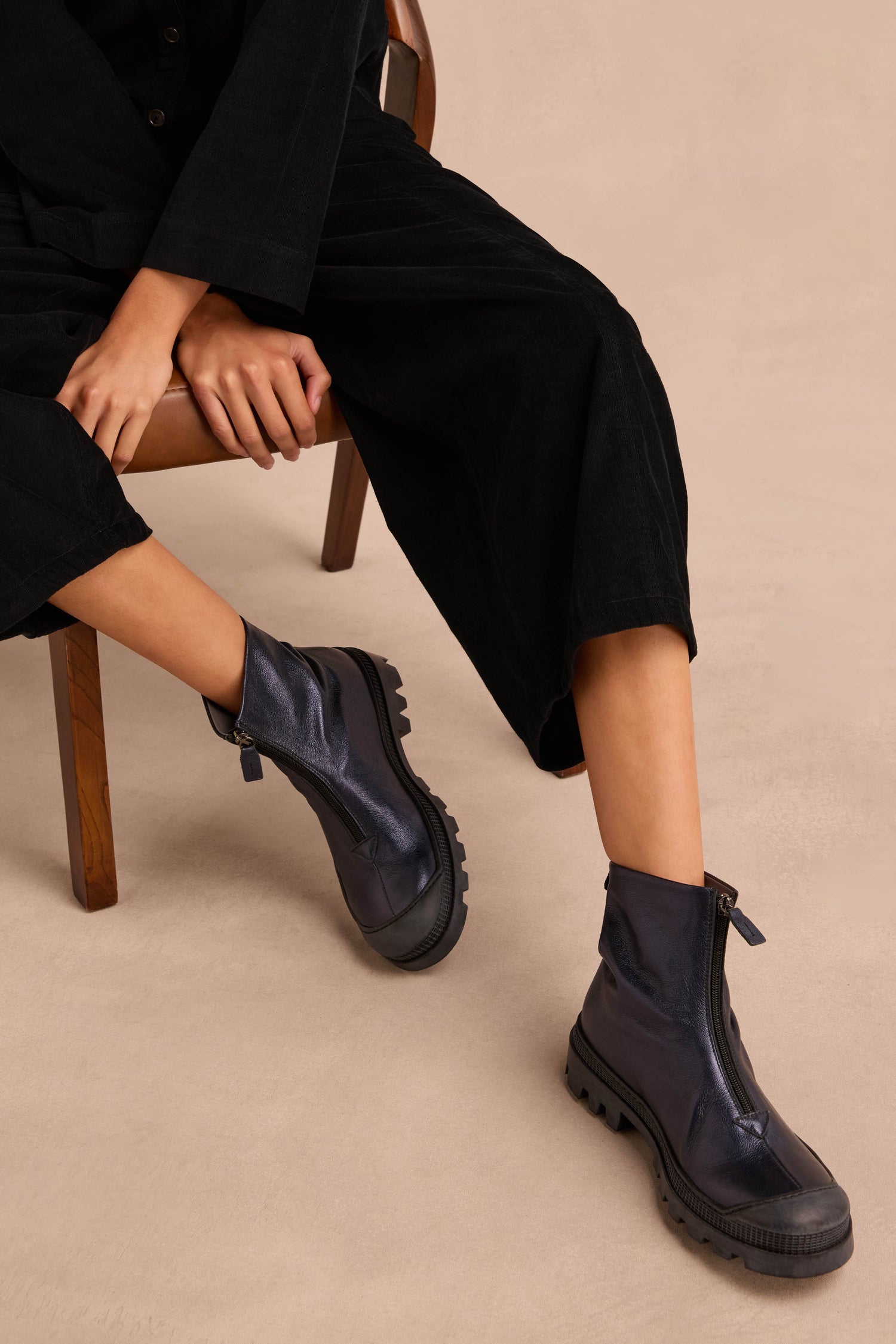 A person seated on a wooden chair, wearing a black outfit and Metallic Blue Violet Zip Front Leather Boots with a chunky tread sole, on a beige background. Their hands rest on their knees.