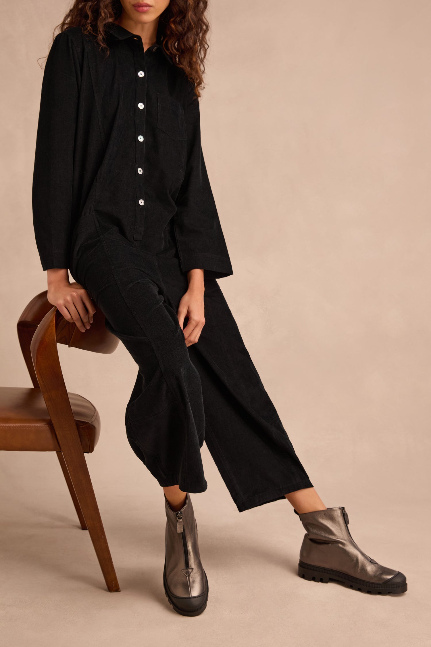 A person is seated on a brown chair against a beige background, wearing a black button-up shirt, black pants, and Metallic Hematite Zip Front Leather Boots featuring a chunky tread sole.