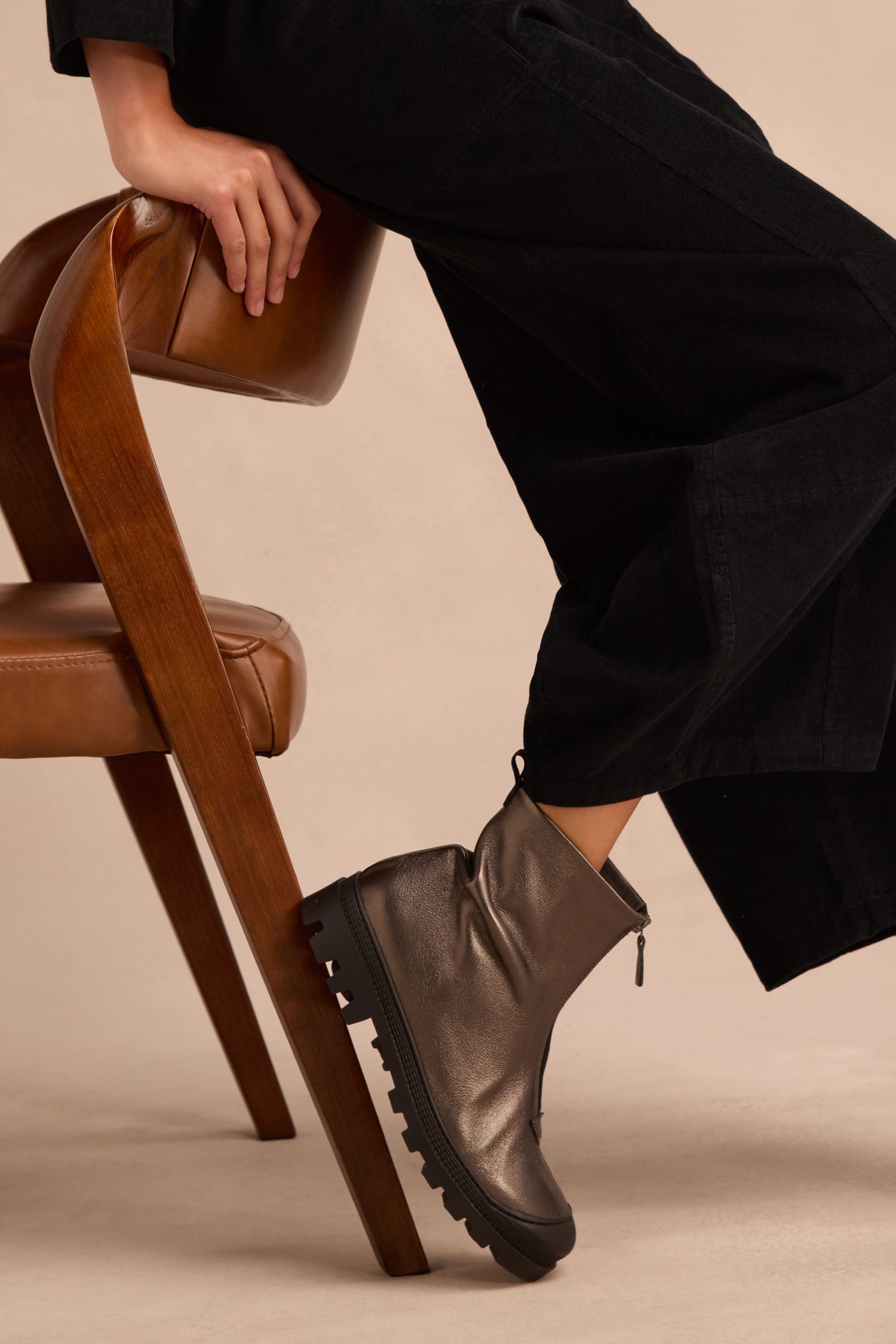 A person is seated on a wooden chair, shown from the lower body, wearing black pants and Metallic Hematite Zip Front Leather Boots with a chunky tread sole. One hand rests on the chair.