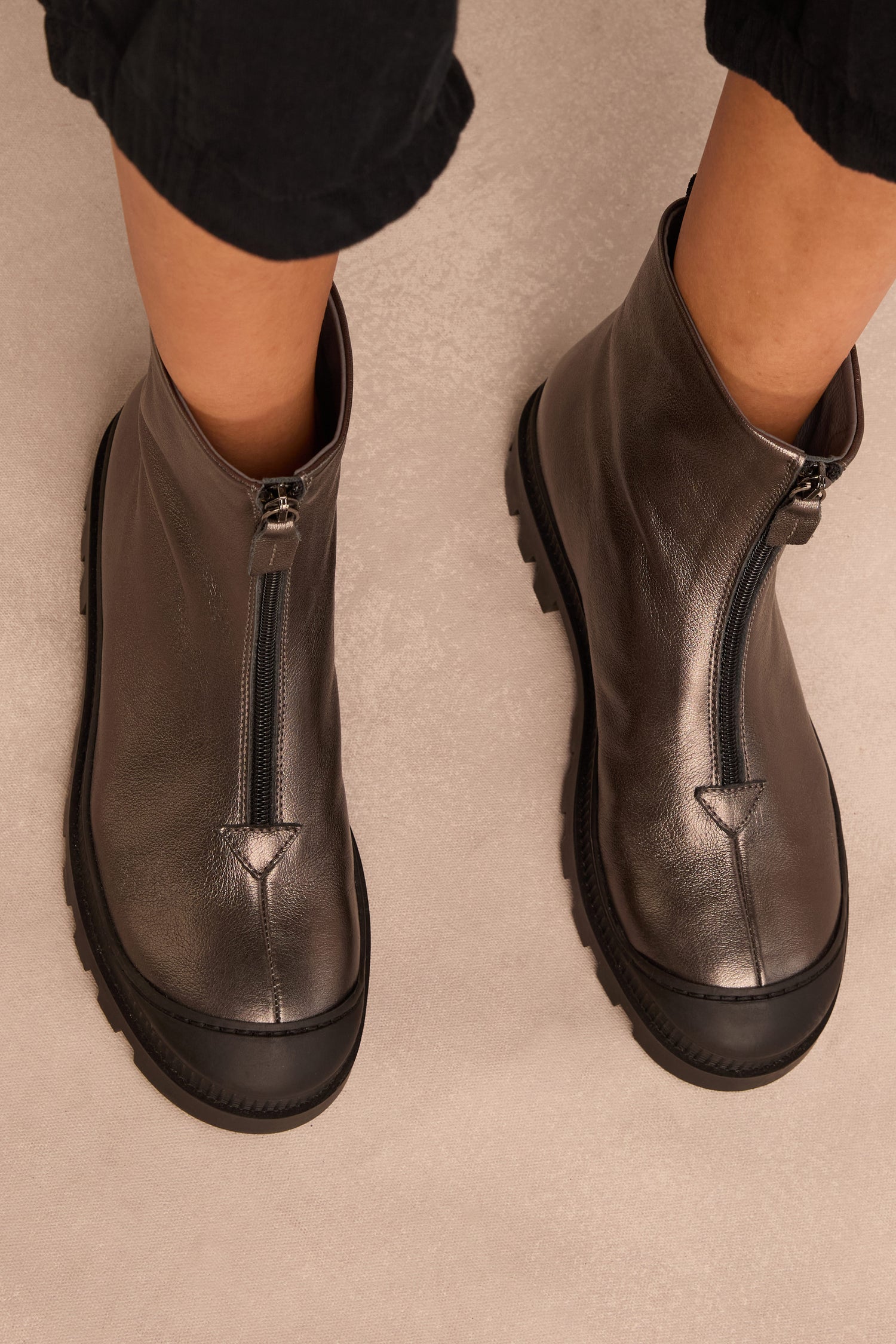 Wearing the Metallic Hematite Zip Front Leather Boots, which feature a chunky tread sole and ankle-high zip front design, the person stands on a light-colored surface.