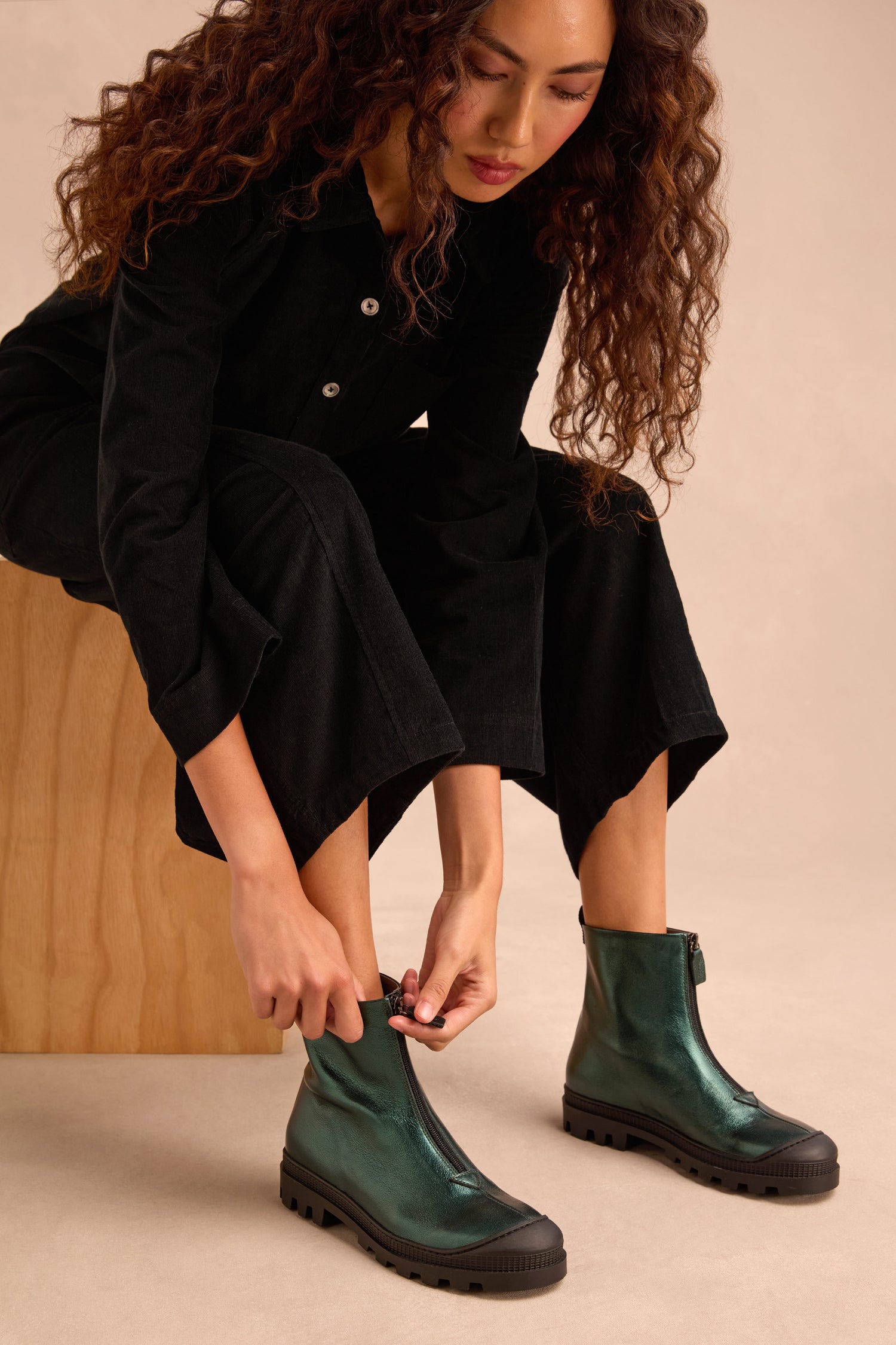 A person with long curly hair sits on a wooden block, dressed in black and putting on Metallic Forest Zip Front Leather Boots with shiny green leather and chunky tread soles.
