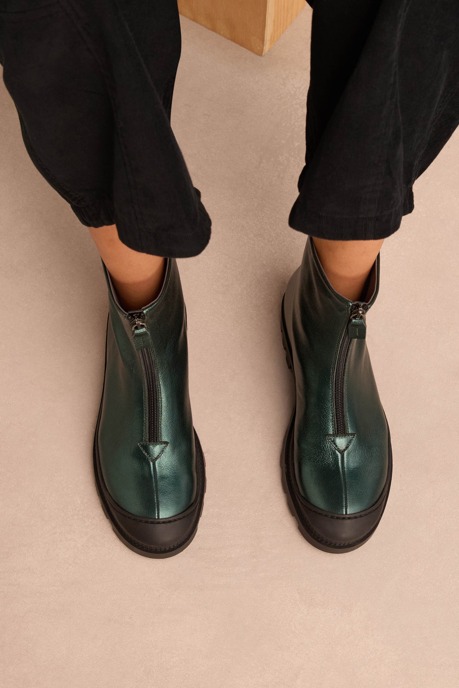 Wearing Metallic Forest Zip Front Leather Boots featuring a chunky tread sole in green, paired with black pants, while standing on a light beige floor.
