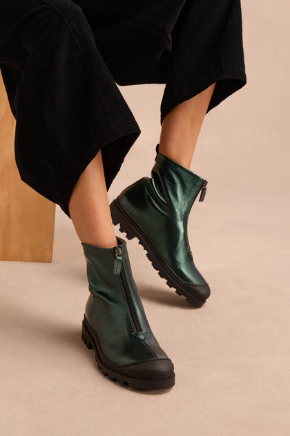 Person wearing loose black pants and Metallic Forest Zip Front Leather Boots with chunky tread soles, sitting on a light-colored surface.