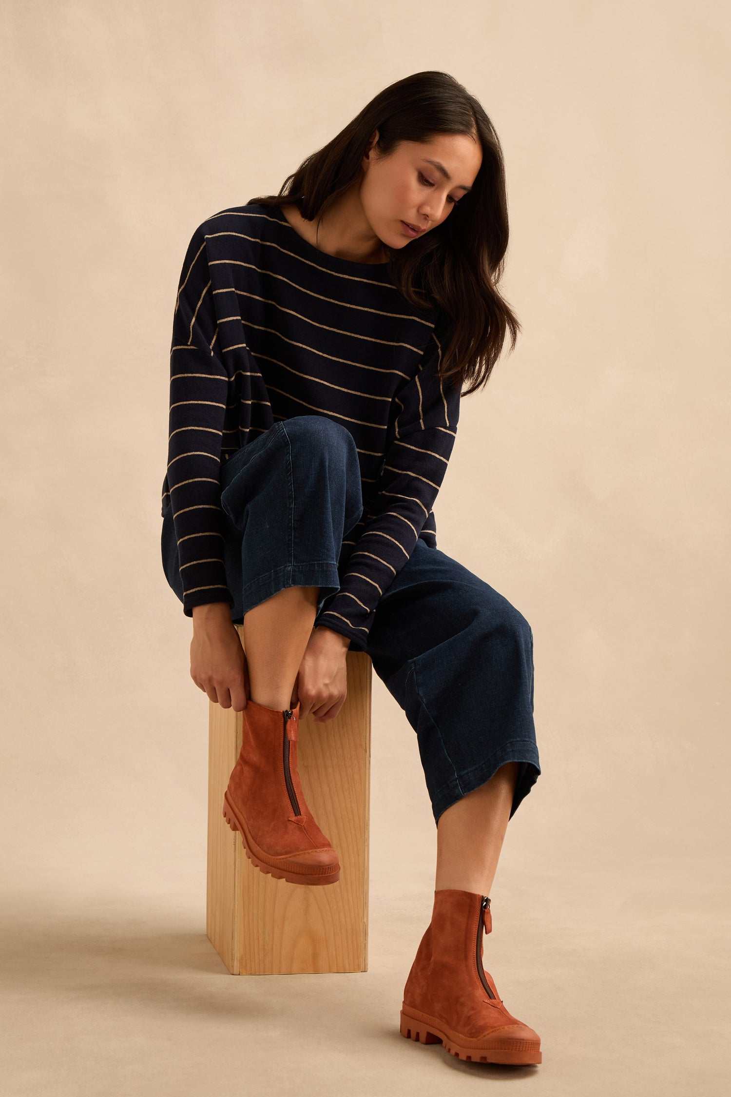 A woman sits on a wooden block, wearing wide-leg denim pants, a navy striped sweater, and the Zip Front Suede Boots—handmade in Italy—looking down as she adjusts her boot.