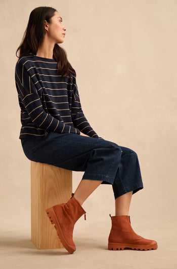 A woman sits on a wooden block, wearing a navy striped long-sleeve shirt, wide-leg denim pants, and brown Zip Front Suede Boots. The boots are handmade in Italy, bringing artisanal craftsmanship to her effortlessly chic style.