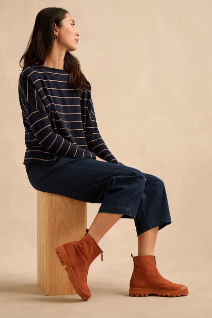 A woman sits on a wooden block, wearing a navy striped long-sleeve shirt, wide-leg denim pants, and brown Zip Front Suede Boots. The boots are handmade in Italy, bringing artisanal craftsmanship to her effortlessly chic style.
