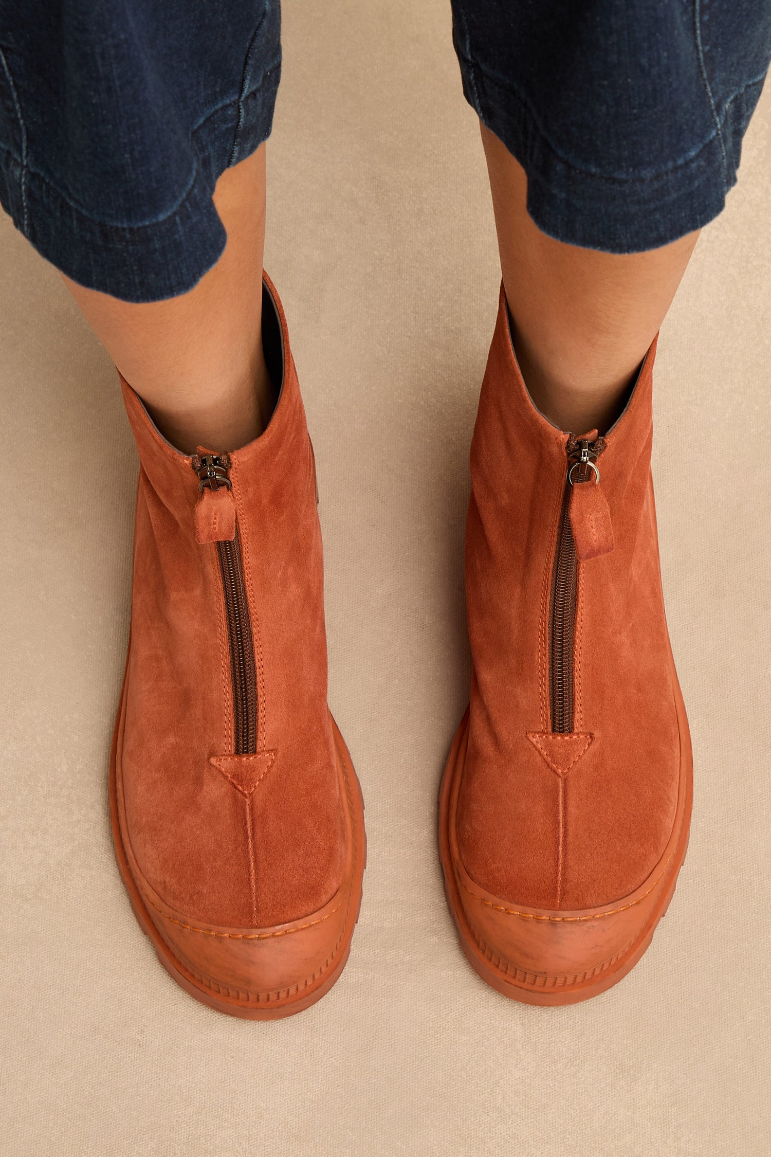Wearing rust-colored Zip Front Suede Boots with front zippers, paired with cropped blue jeans and standing on a light beige surface. These stylish ankle boots are handmade in Italy.