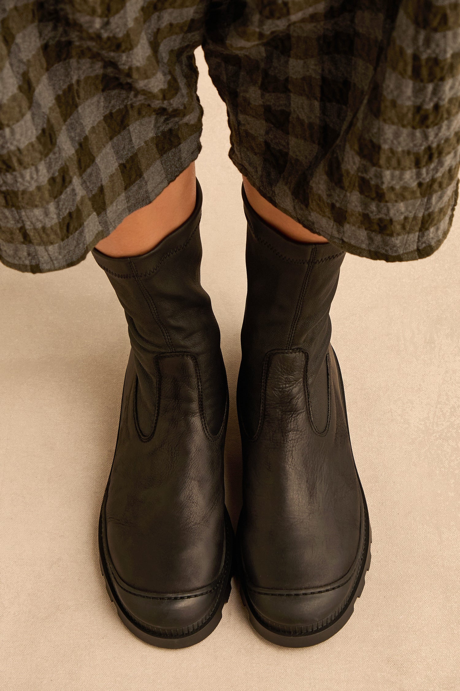 Person wearing Stitch Detail Napa Leather Stretch Boots in black and a plaid skirt, standing on a beige surface.