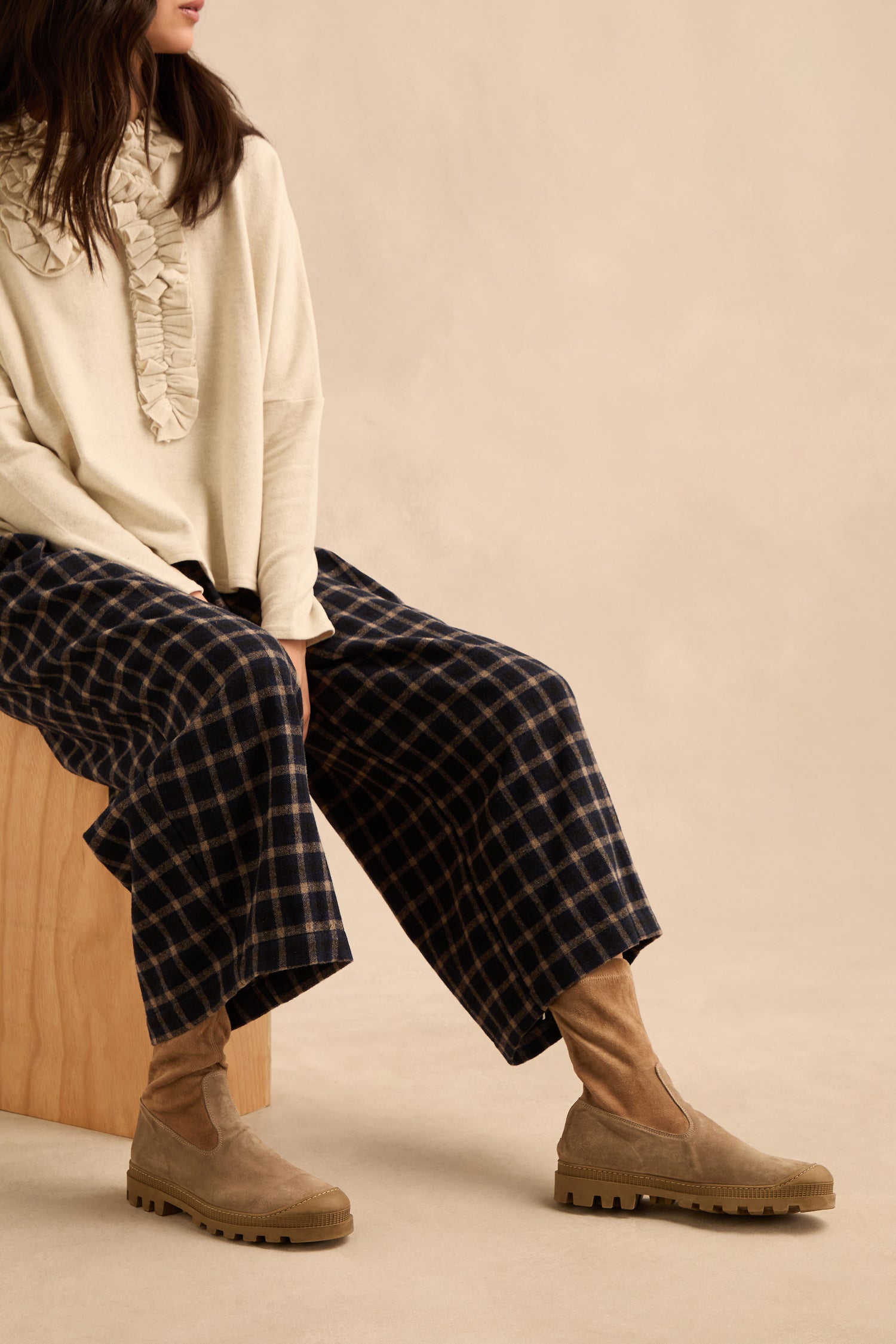 A woman sits on a wooden block against a neutral background, wearing a cream ruffled sweater, plaid wide-leg pants, and Stitch Detail Suede Stretch Boots—beige ankle boots handmade in Italy.