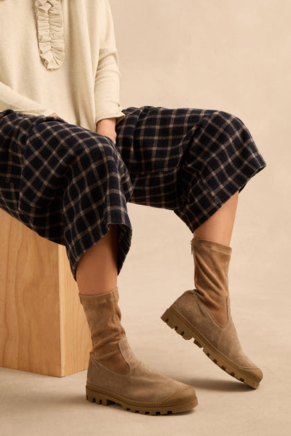 Person sitting on a wooden block wearing a cream top, plaid navy culottes, and Stitch Detail Suede Stretch Boots handmade in Italy from tan suede with rugged soles.