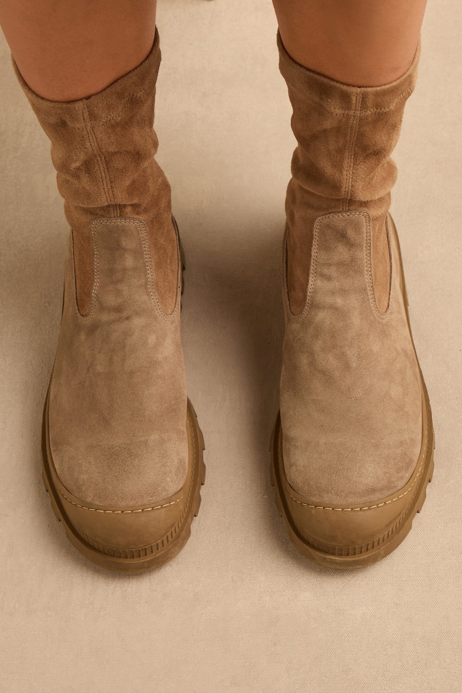 A person wearing the Stitch Detail Suede Stretch Boots, featuring thick soles, stands on a light-colored floor.
