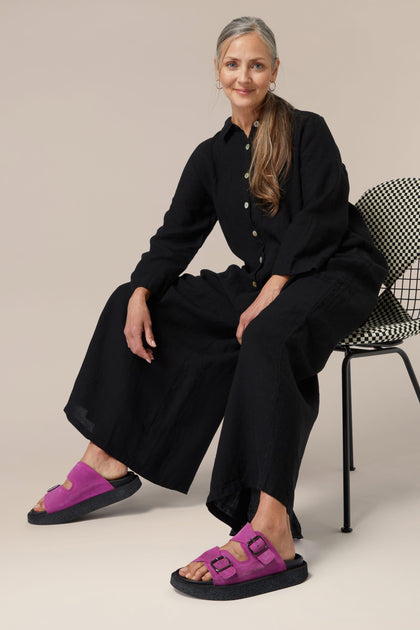 Woman seated on a chair wearing a black jumpsuit and Italian construction purple Suede Double Buckle Slider sandals.