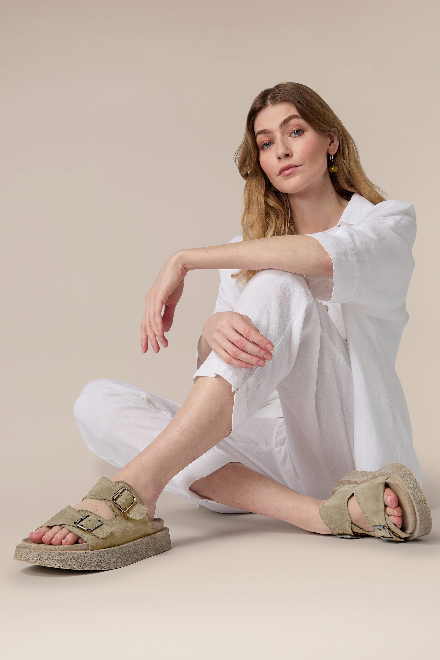 A woman sitting on the floor, casually dressed in a white outfit and wearing Italian construction Suede Double Buckle Slider sandals, with a neutral backdrop behind her.