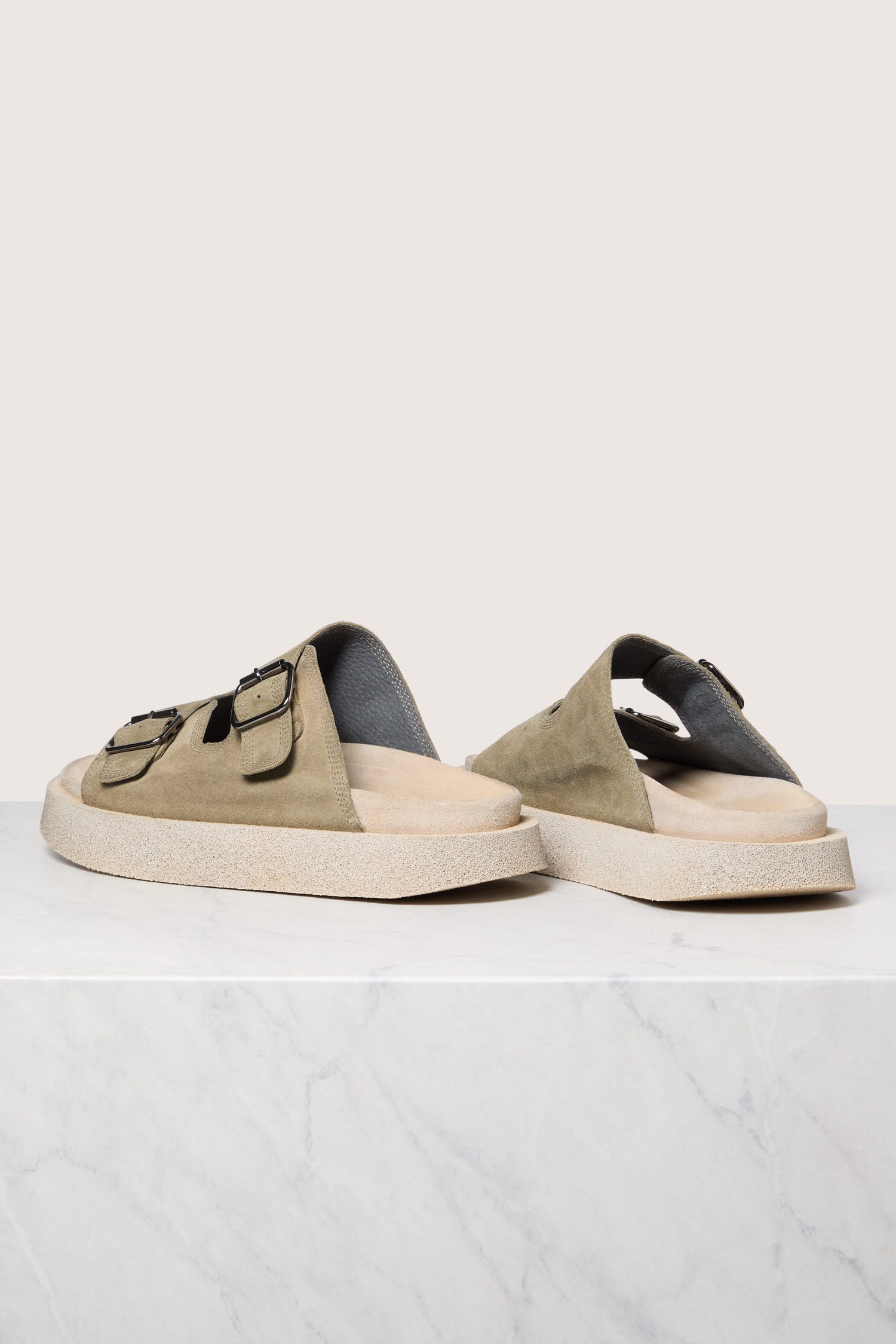 A pair of beige Suede Double Buckle Sliders on a white surface with a marble backdrop.