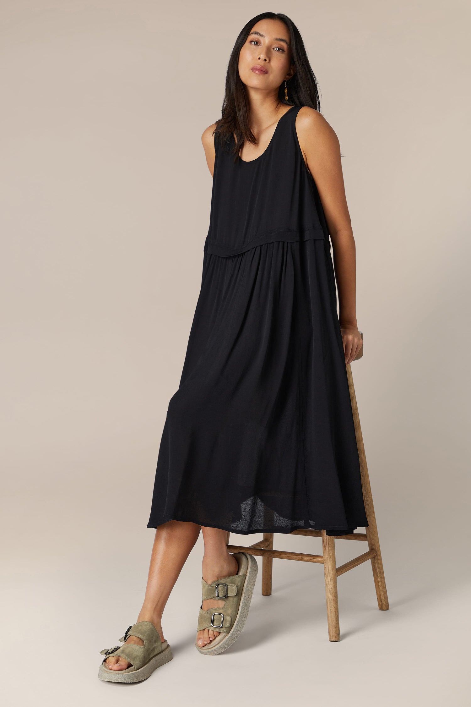 Sentence with replaced product name: A woman standing against a neutral background wearing a sleeveless black midi dress and Suede Double Buckle Sliders.